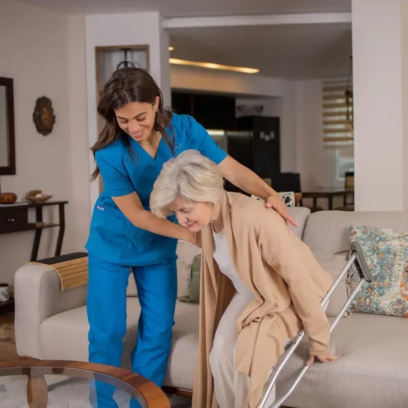 A caregiver in blue scrubs assists an older woman using crutches to stand up from a sofa in a cozy living room, conveying care and support.