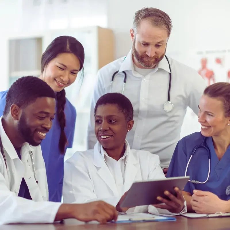 A diverse medical team, including men and women in scrubs and white coats, gathers around a tablet, smiling and engaged in discussion.