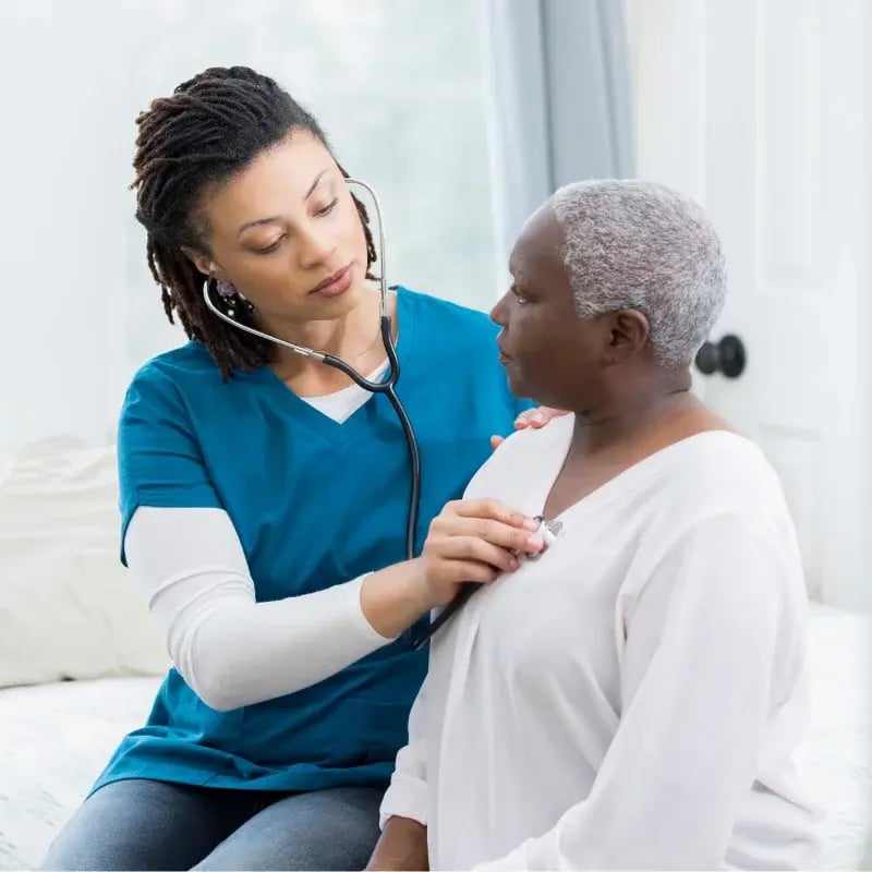 A female healthcare professional in blue scrubs uses a stethoscope to check an older woman’s heartbeat in a softly lit room.