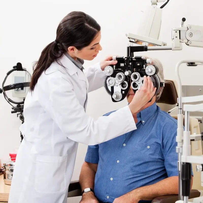 A female optometrist in a white coat adjusts a phoropter on an older mans face during an eye exam in a clinic