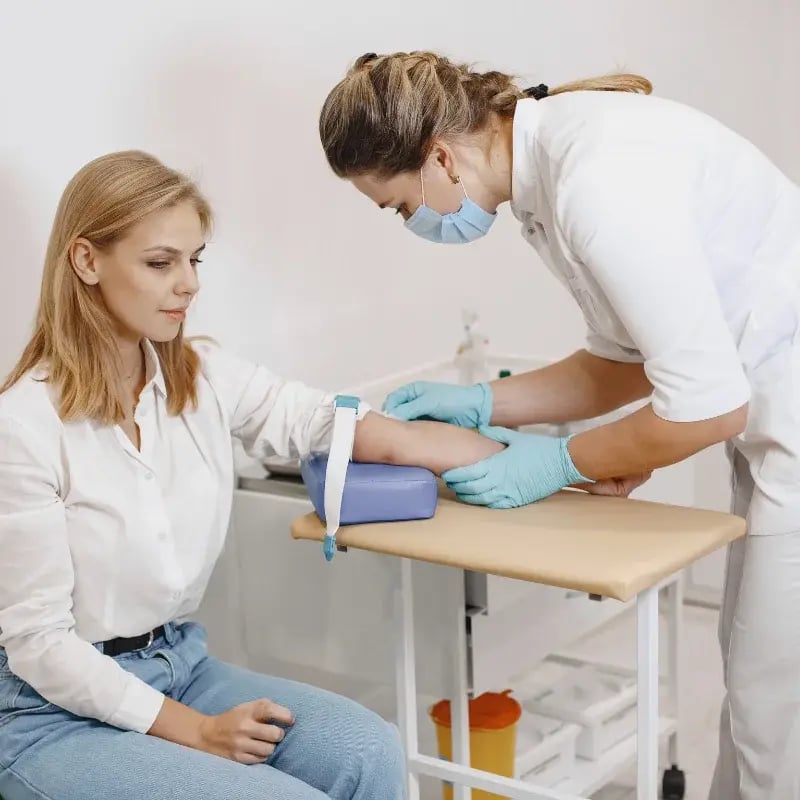A female phlebotomist, wearing a mask and gloves, prepares to draw blood from a seated woman in a clinic