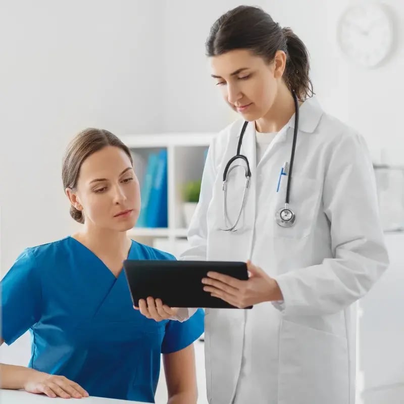 A female physician in a white coat, holding a tablet, talking with a female nurse in blue scrubs at a medical office.