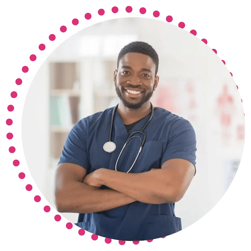 A male nurse in navy scrubs stands confidently with arms crossed, a stethoscope hanging from his neck in a hospital environment.