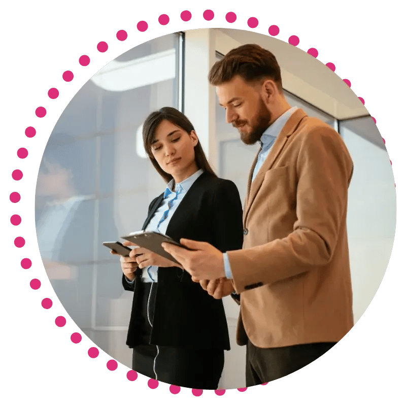 A man and woman in business attire stand in a modern office, reviewing documents together. A pink dotted circle frames them.