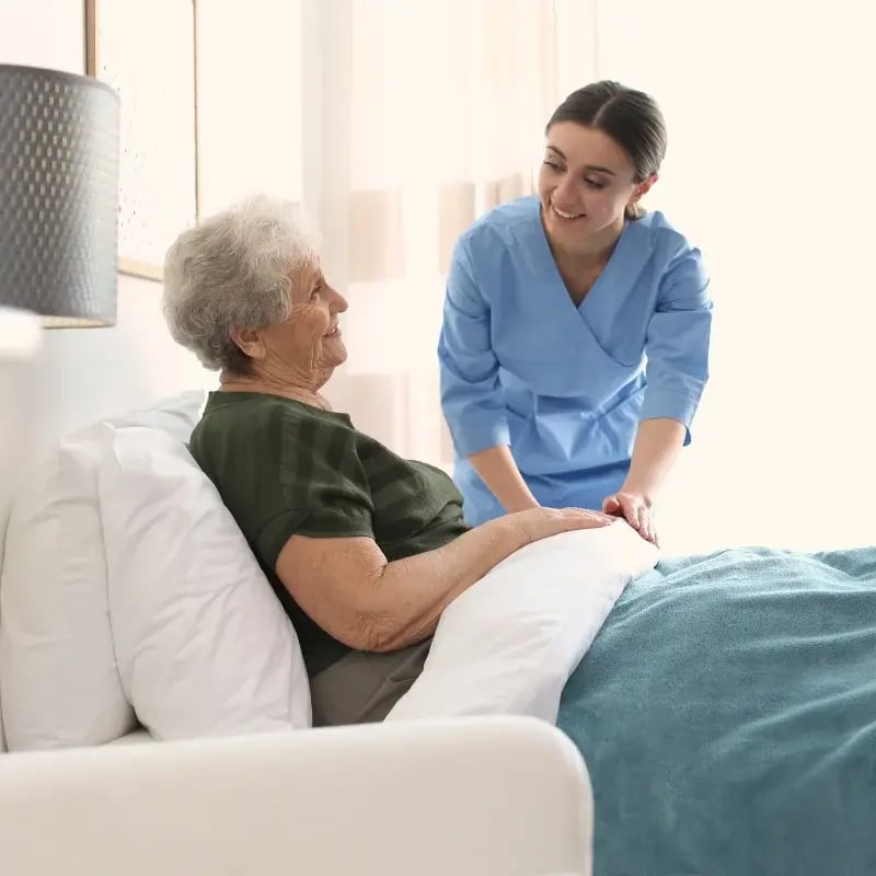 A nurse in blue scrubs kindly assists a smiling elderly woman sitting up in bed.