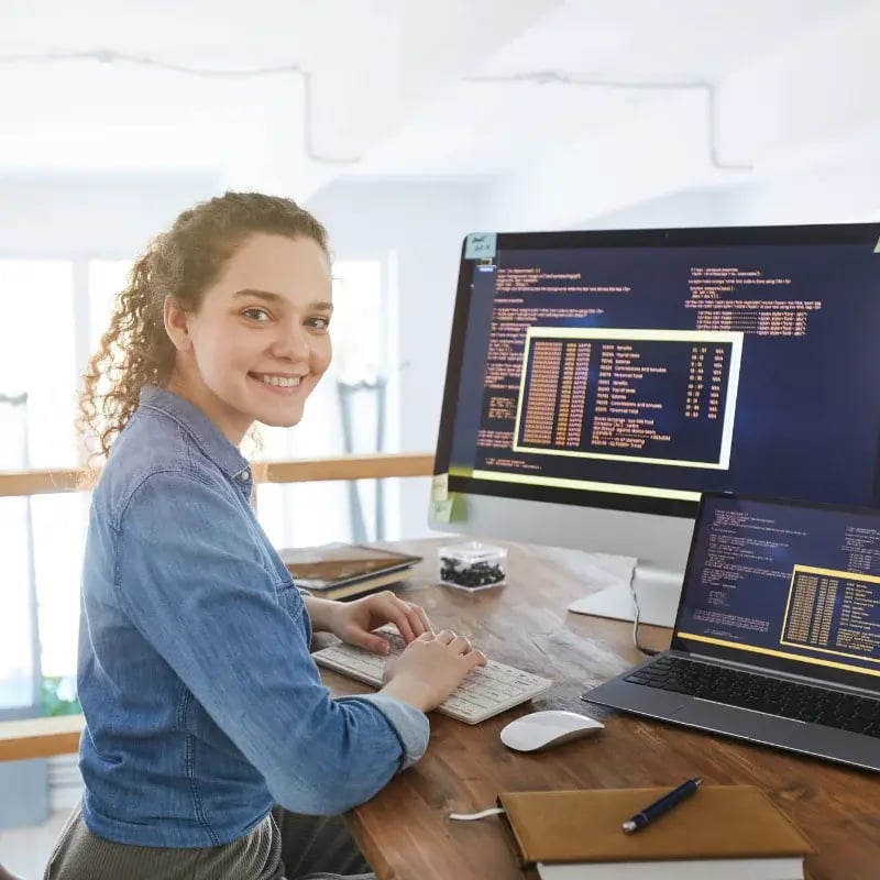 A person sits at a wooden desk, coding on dual monitors displaying healthcare programming data, surrounded by notebooks and a computer mouse.