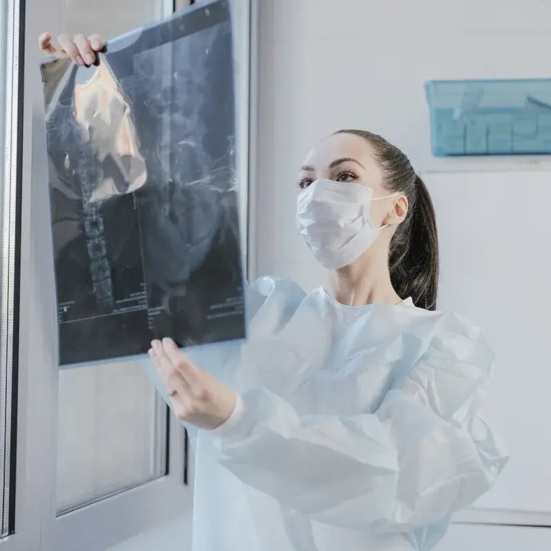 A rad tech in a mask and gown examines an X-ray in a hospital setting