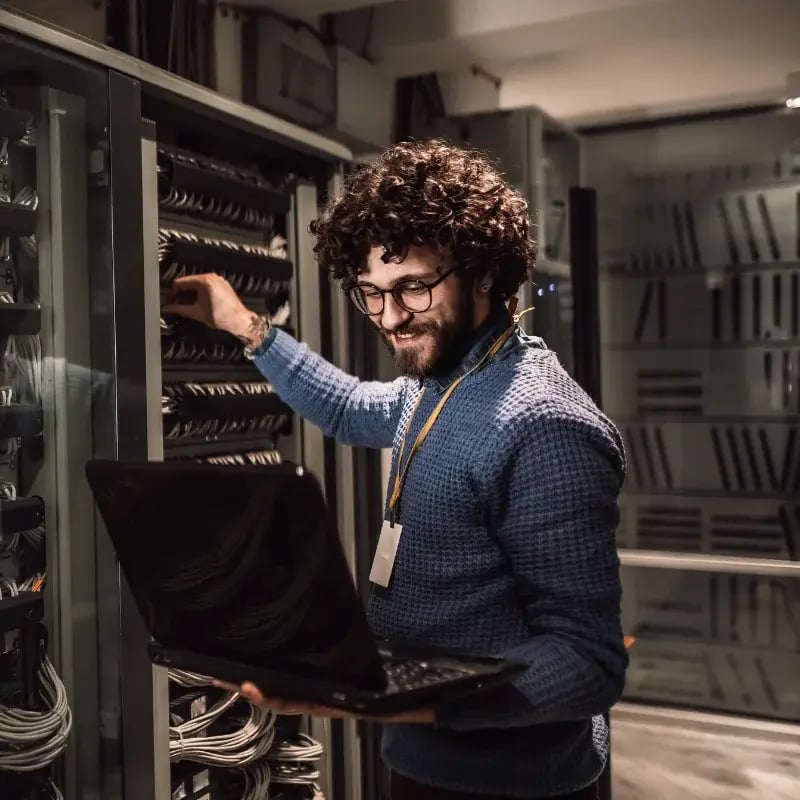 A smiling man with curly hair and glasses works in a server room, holding a laptop and adjusting cables in a rack.