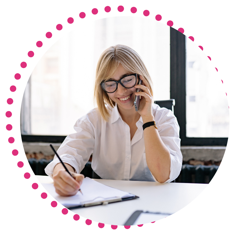 A woman with glasses and a white shirt smiles while talking on the phone. She is writing in a notebook.