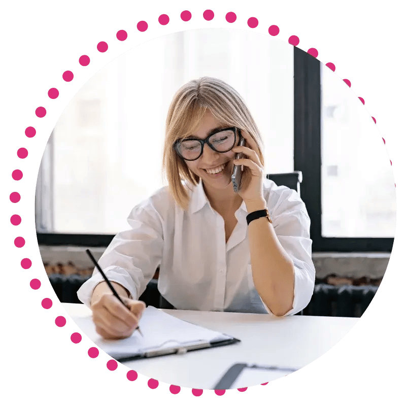 A woman with glasses and a white shirt smiles while talking on the phone. She is writing in a notebook.