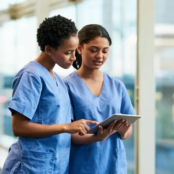 A pair of nurses checking a tablet in a hospital aisle