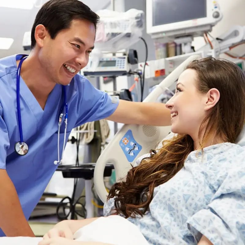 Smiling nurse in blue scrubs interacts with a female patient in a hospital bed.