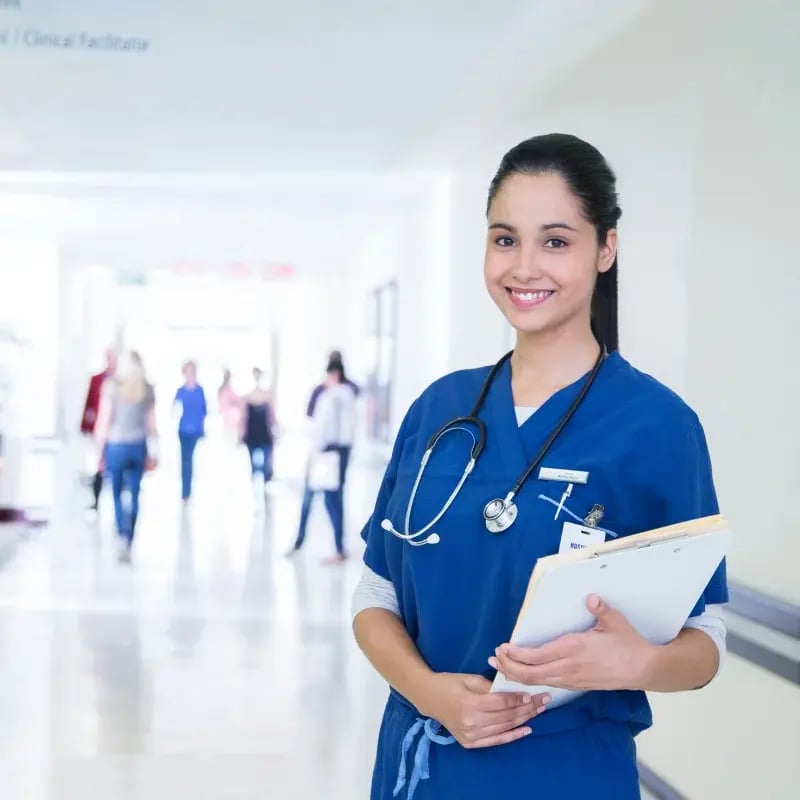 Smiling nurse in blue scrubs with a stethoscope and clipboard in a hospital corridor.