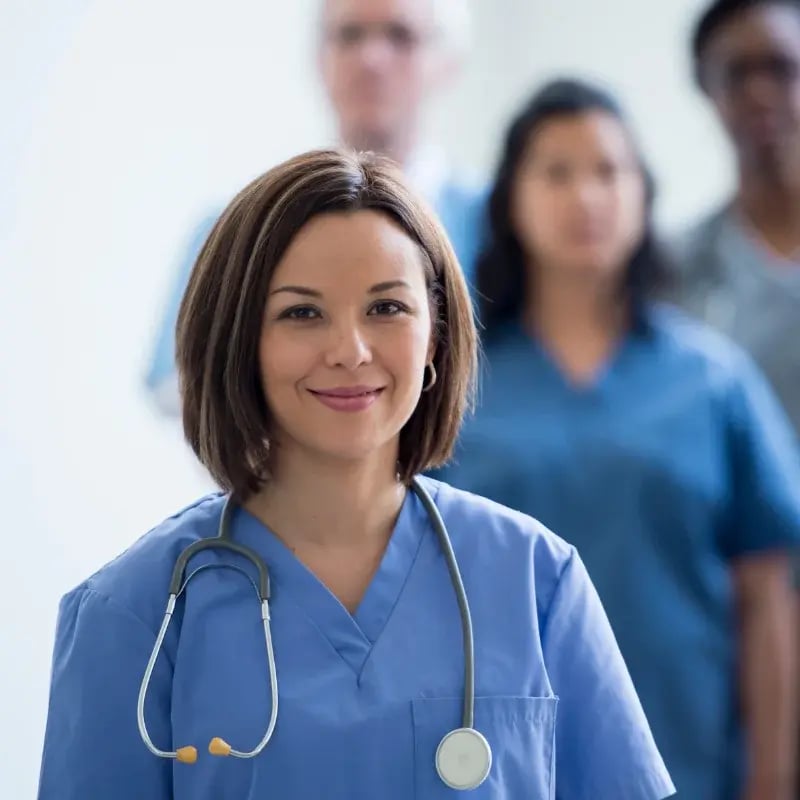 Smiling nurse in blue scrubs with a stethoscope stands confidently in focus, with three blurred healthcare professionals in the background.