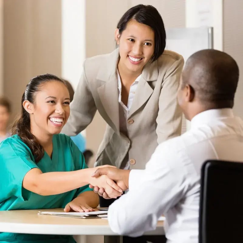 Smiling woman in scrubs shakes hands with a seated man, while another woman in a suit stands beside them, all appearing pleased and professional.