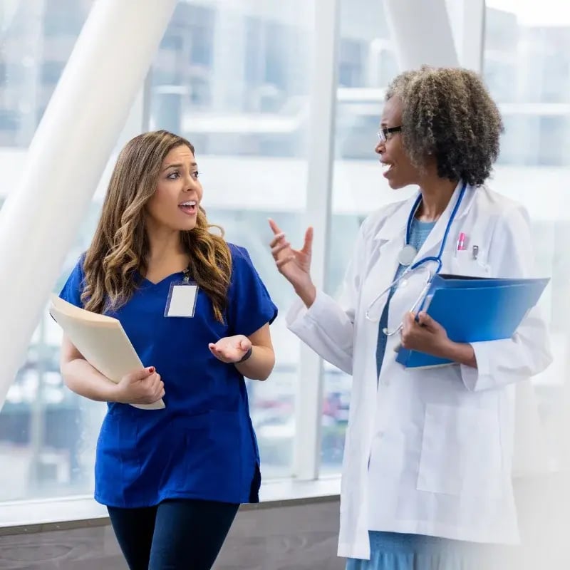 Two healthcare professionals, one in a blue scrub and one in a white coat, are talking in a bright hospital hallway