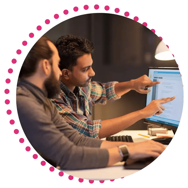Two individuals collaborate at a computer, one pointing at the screen while discussing information in a dimly lit workspace.