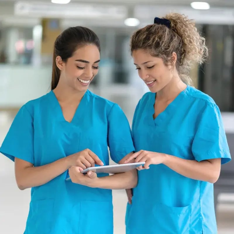 Two smiling healthcare professionals in blue scrubs stand in a hospital, engaging with a tablet. They appear collaborative and focused.
