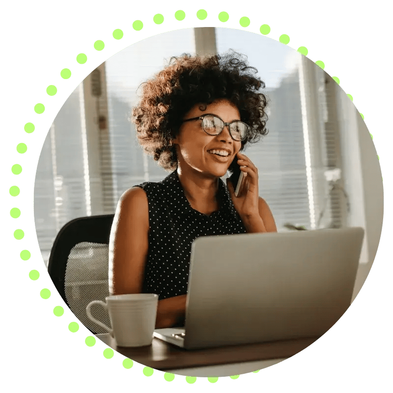A woman smiles while talking on the phone and working on a laptop at a desk with a coffee mug, in an office setting.