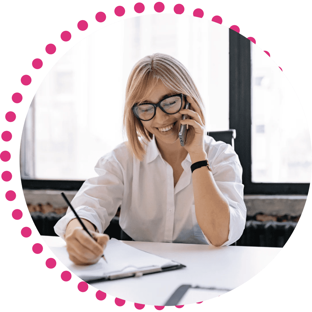 A woman with glasses and a white shirt smiles while talking on the phone. She is writing in a notebook.