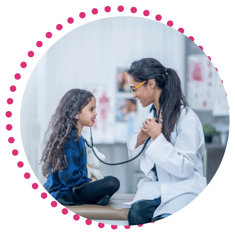 A female doctor smiles warmly at a young girl, who is listening to a stethoscope. They sit together in a light, cheerful medical office.