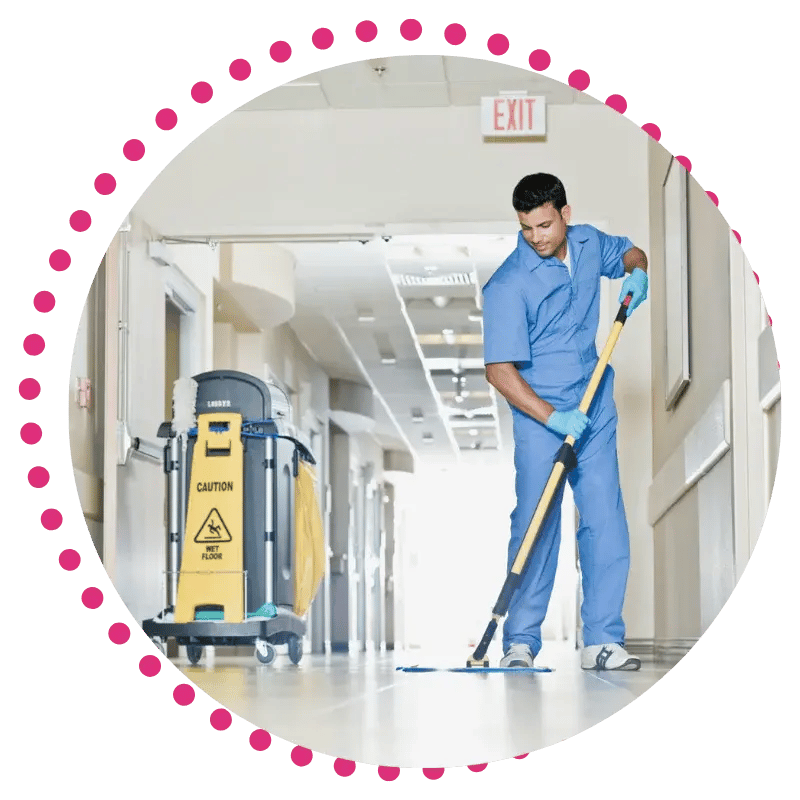 A person in blue scrubs mops a bright, clean hospital corridor. Nearby, a caution sign and cleaning cart emphasize a tidy, professional environment.