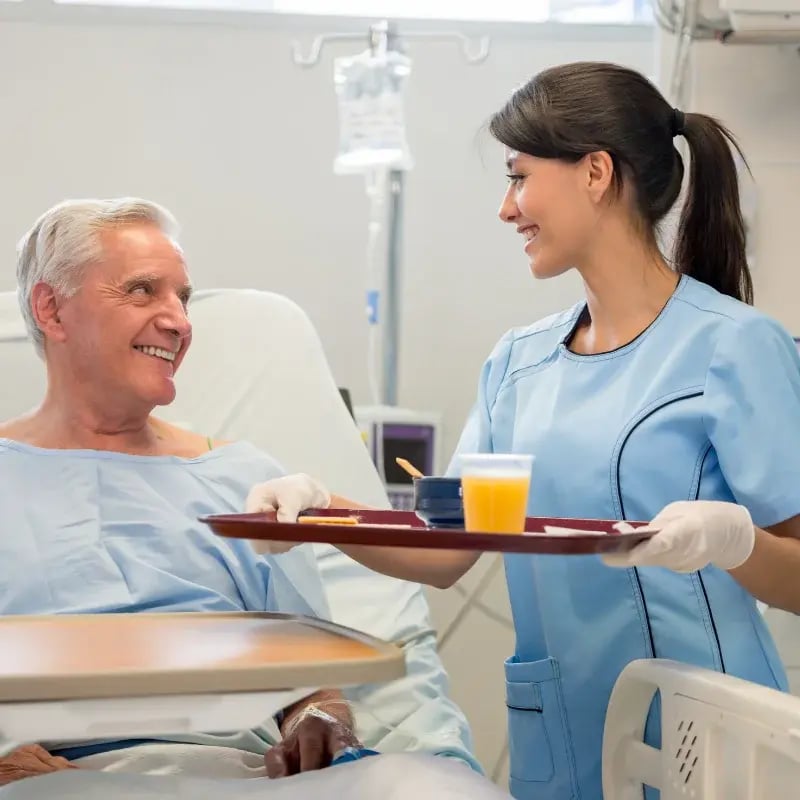 A nurse in blue scrubs smiles while serving a hospital patient a tray with food and juice. The patient, in a gown, sits in bed, smiling back warmly.
