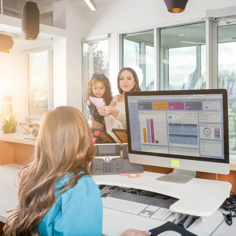 A woman and a child interact with a receptionist in a bright, modern office. The woman hands over paperwork; a computer monitor displays charts.