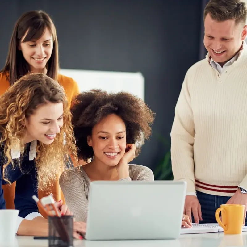A diverse group of people gathered around a laptop, engaged in discussion and looking at the screen together.