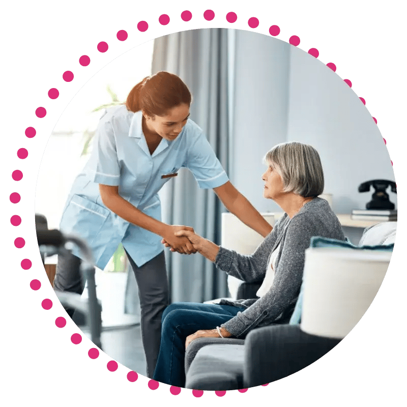 A nurse in uniform smiles warmly while helping an elderly woman stand from a couch in a cozy living room