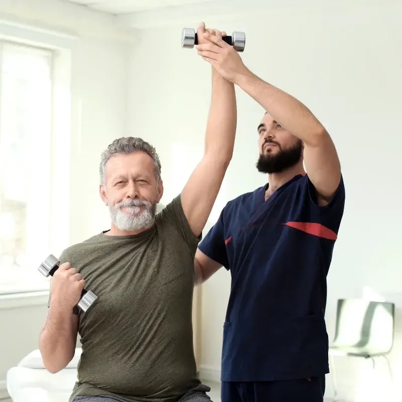 A bearded man lifts a dumbbell with help from a therapist in a bright therapy room.